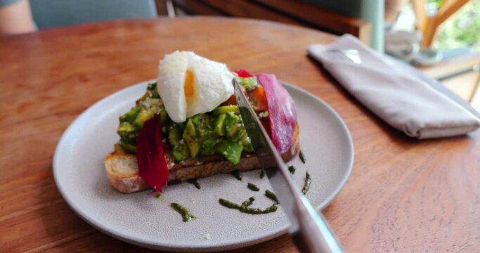 Close-up view from the table knife perspective, it approaches and cuts a poached egg on top of avocado toast. Unusual angle shot of starting eating breakfast meal