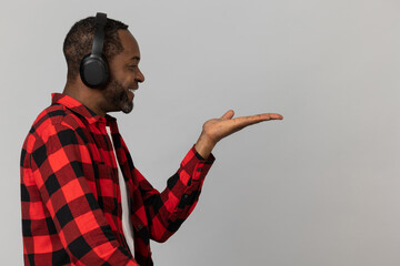 Smiling black bearded man in headphones wearing red checkered shirt presenting copy space on palm