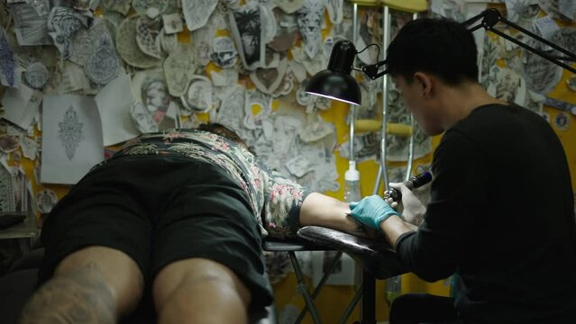 Asian tattoo artist making a tattoo to a lied down customer in a studio