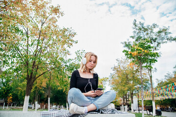 A cute teenage girl with a phone in her hand is sitting on a bench in a modern park. A schoolgirl writes messages to friends on the Internet. Social networking, listening to music with headphones.