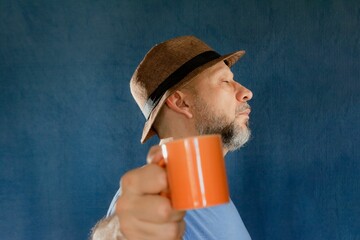 Man wearing a wide-brimmed hat and holding a bright orange mug filled posing in a studio