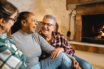 Multiracial senior friends by wood fireplace having fun together at rural home. Winter and fall...