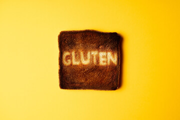 Slice of white toast bread burnt on toaster with word Gluten on it. Bright yellow background. gluten intolerance concept - obrazy, fototapety, plakaty