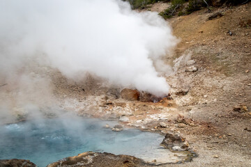 small geyser in yellowstone park national park, image shows a small gyser and a crystal clear blue heated pool of water, with sand and sulphur surrounding, taken october 2023