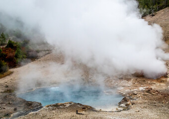 small geyser in yellowstone park national park, image shows a small gyser and a crystal clear blue heated pool of water, with sand and sulphur surrounding, taken october 2023