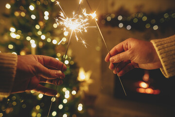 Happy New Year! Burning sparklers in hands  in eve on background of modern fireplace and christmas tree with golden lights. Fireworks glowing in hands, couple celebrating in festive dark room