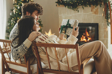 Happy couple in cozy sweaters exchanging stylish wrapped christmas gifts on background of fireplace...