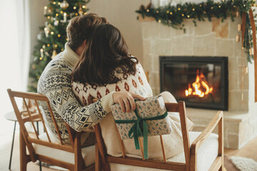 Happy young family in cozy sweaters exchanging stylish christmas gifts on background of fireplace...