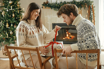 Happy couple in cozy sweaters opening stylish wrapped christmas gifts on background of fireplace with festive mantle and modern decorated christmas tree with lights. Merry Christmas!