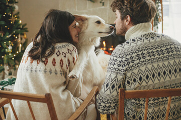 Happy young family in cozy sweaters hugging with cute dog and relaxing on background of fireplace...