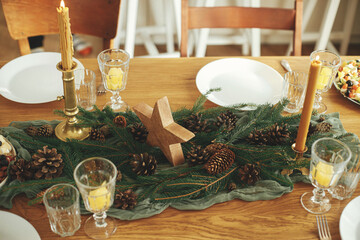 Christmas rustic table setting. Stylish table runner with fir branches and pine cones, candles, cheese appetizers and starters, glasses and cutlery. Holiday dinner arrangement. Happy new year!