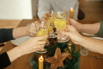 Hands with mimosa cocktail glasses clinking on background of stylish christmas table setting with fir branches, pine cones and candles. Friends and family celebrating and toasting, happy holidays
