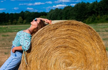 Naklejka na ściany i meble Young woman in blue jeans and sunglasses standing against a round hay bale in a rural area