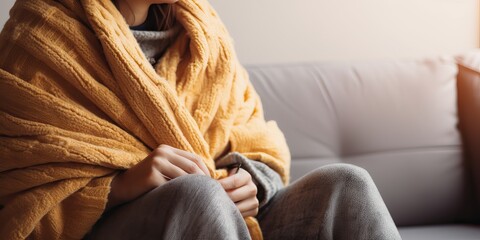 It's cold in the house in winter. Person freezing, fever or trouble with heating. Sad person in wool plaid and scarf and wearing warm hat sitting on sofa at home in wintertime - obrazy, fototapety, plakaty