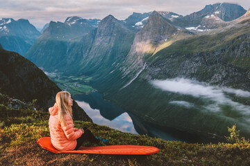 Woman traveling in Norway alone enjoying mountains and fjord view sitting on sleeping pad camping...