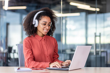 Young beautiful woman inside the office works with a laptop, a businesswoman in headphones listens...