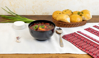 A plate of borsch and pampushka buns with garlic and herbs on a Ukrainian-style table with an embroidered tablecloth. Traditional Ukrainian borsch.