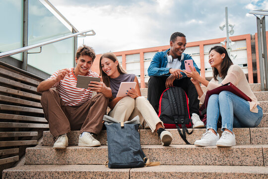 Group of multiracial high school students talking on a staircase at university campus, using a tablet app and smartphone to do homework project after class. Friendly teenage people studying together