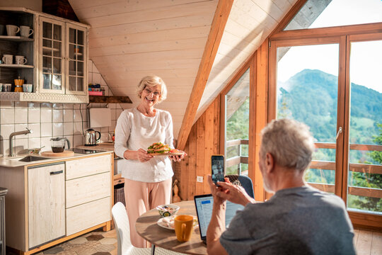 Happy senior woman holding plate with sandwich with husband taking picture in wood cabin