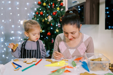 Little girl watching her mother painting a pattern with a toothpick on a glazed cookie