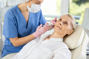 Elderly woman getting injections of hyaluronic acid filler in nasolabial folds for skin tightening...
