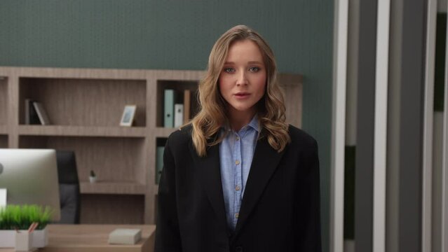 Attractive young blonde with short curls, blue eyes in formal outfit standing in modern office looking at camera with effect of moving away from camera. High quality 4k footage