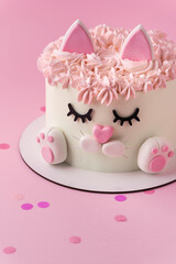 Cute birthday cake in the shape of cat with mastic ears and paws decorated with pink cream fur on the pink background. Adorable cake for a little girl who likes cats