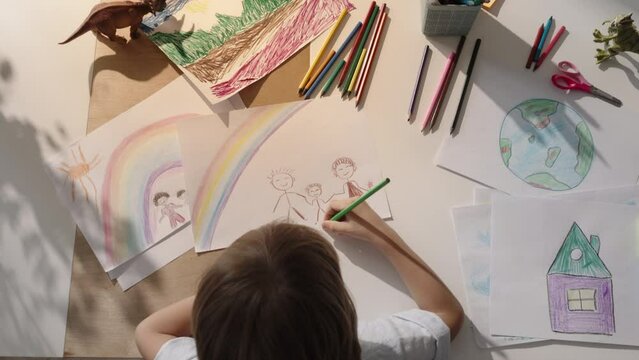 Top view of boy at his desk. Painting family under bright rainbow. Drawing of happy family. Children's creativity, children's psychological test for child's perception of family relationships. 