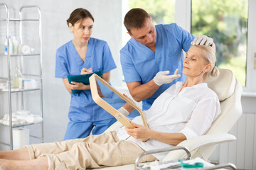 Beautician talking with patient before injection treatment, while client observing herself in...