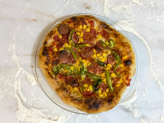 Traditional Italian pizza with corn, peppers and sausage on a white marble floor scattered with flour. Selective focus, close up. 