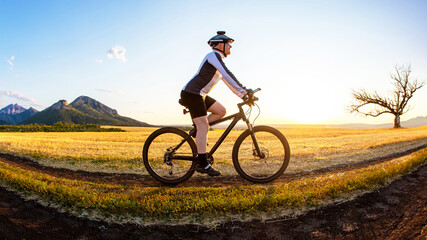 The cyclist rides a bike on the road near the field against the backdrop of the setting sun. Outdoor sports. Healthy lifestyle.