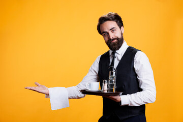 Skilled waiter shows direction to table at restaurant, serving people as occupation. Young man working as butler with meal tray in hand, pretends to serve cup of coffee to customers on camera. - Powered by Adobe