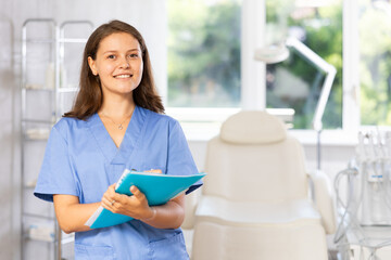 Young female cosmetologist in medical uniform taking notes in folder in medical room