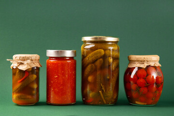 Canned vegetables in jars on a green background. Pickled cucumbers, tomatoes, chili sauce