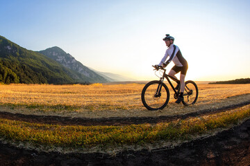 The cyclist rides a bike on the road near the field against the backdrop of the setting sun....