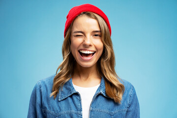 Closeup portrait smiling American  woman winks wearing stylish red hat isolated on blue background. Happy modern hipster with stylish hair looking at camera. Positive lifestyle concept - obrazy, fototapety, plakaty