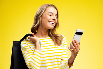 Attractive smiling woman holding smartphone and black shopping bags using mobile app shopping online isolated on yellow background. Black Friday, sale, internet store concept
