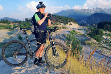 The cyclist with the Bicycle and binoculars in hand stands on a sandy hill in the sunlight.