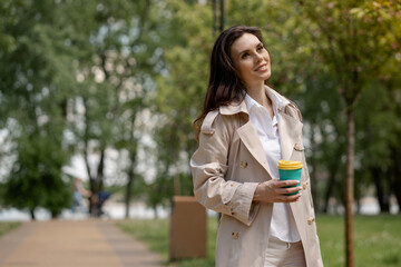 A cute girl brunette walks in the spring park and holds a cup of coffee in hands. A smiling lady...