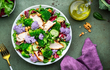Healthy fresh salad from purple cauliflower, broccoli, grilled chicken breast, cucumbers, red onion, radicchio, spinach with walnuts, green table background, top view