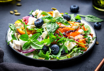 Tasty salad with grilled peaches, blueberries, onion, feta cheese, basil and arugula on black table background, top view