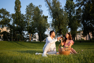 Romantic caucasian couple drinking red wine at urban park background. Happy family enjoying picnic and drinks alcohol in glasses. Love story in nature. Cute boyfriend with girlfriend resting on