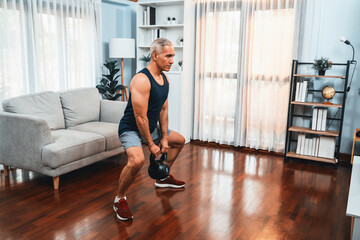 Athletic and sporty senior man engaging in leg day training session with squat and bodyweight kettle ball at home exercise as concept of healthy fit body lifestyle after retirement. Clout - obrazy, fototapety, plakaty