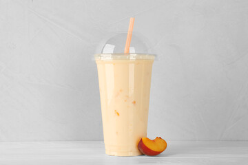 Plastic cup of tasty smoothie and fresh peach on white wooden table