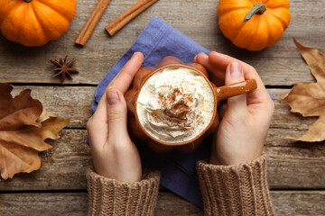 Woman holding mug of pumpkin spice latte with whipped cream at wooden table, top view