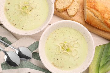 Bowls of tasty leek soup, spoons and bread on white wooden table, flat lay