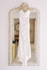 Wedding dress with a train hangs on a hanger on an antique mirror in a carved frame
