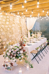 Bouquets of flowers and lit candles stand on a festive table on the terrace in the light of garlands