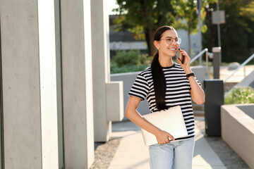 Happy young woman with modern laptop and talking on smartphone outdoors. Space for text