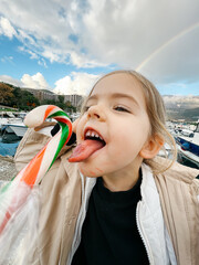 Little girl with her tongue hanging out reaches for a lollipop against the backdrop of a rainbow